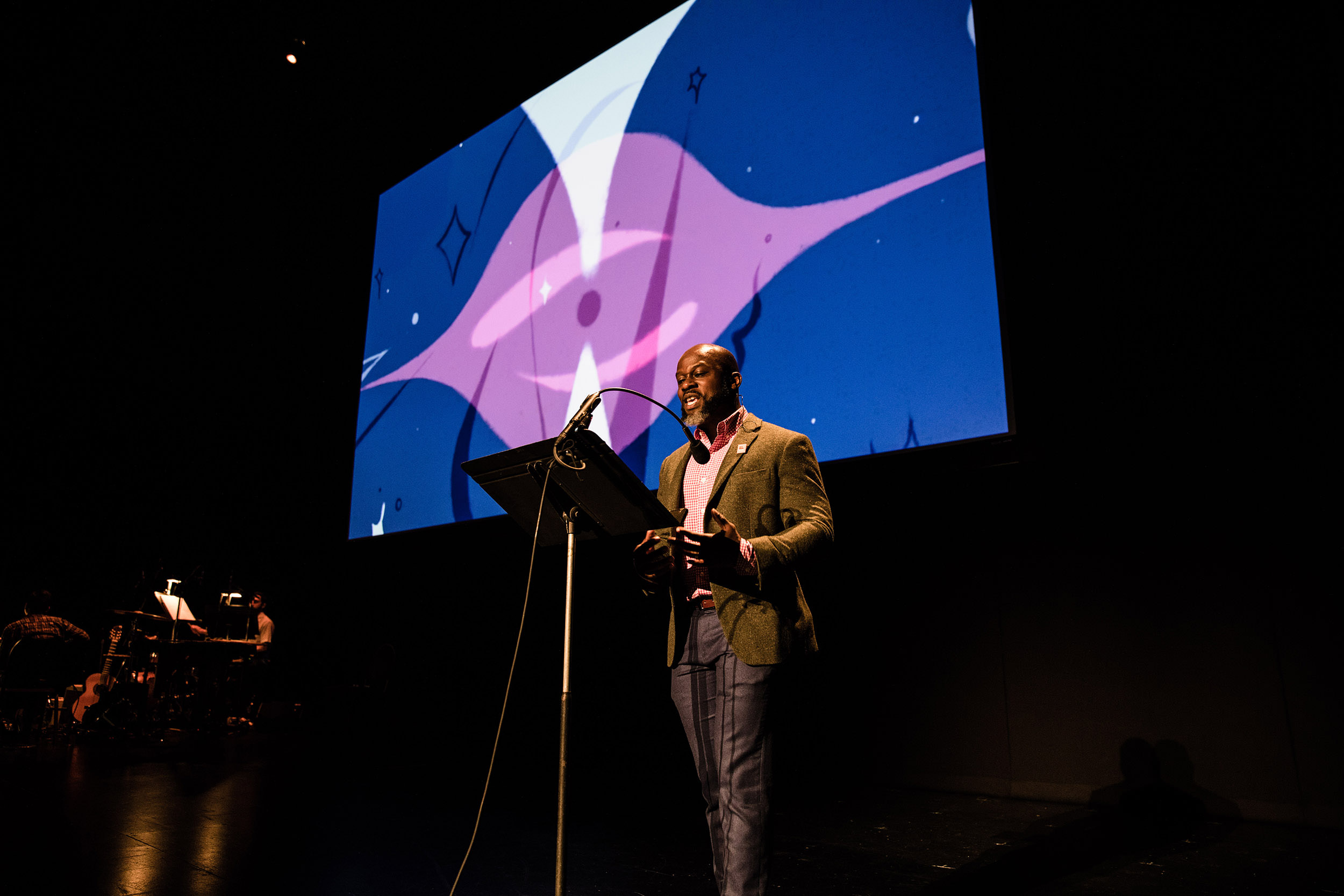 Marc Bamuthi Joseph performs on stage in front of a screen with blue and purple artwork.