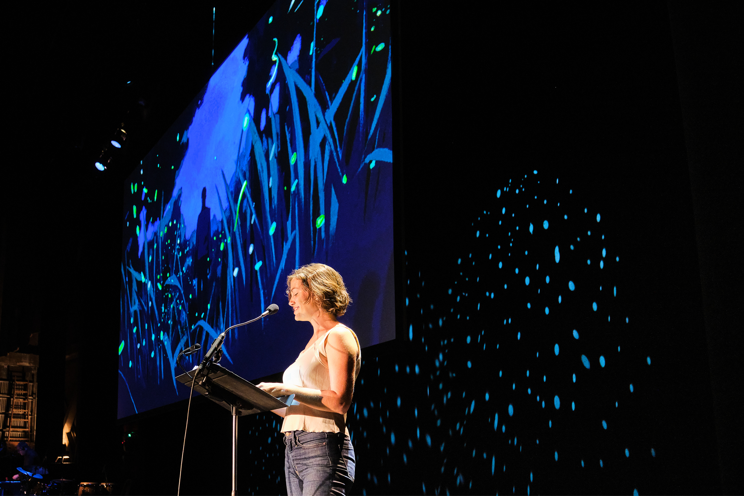 Brooke Jarvis performs on stage in front of a screen with artwork of fireflies.