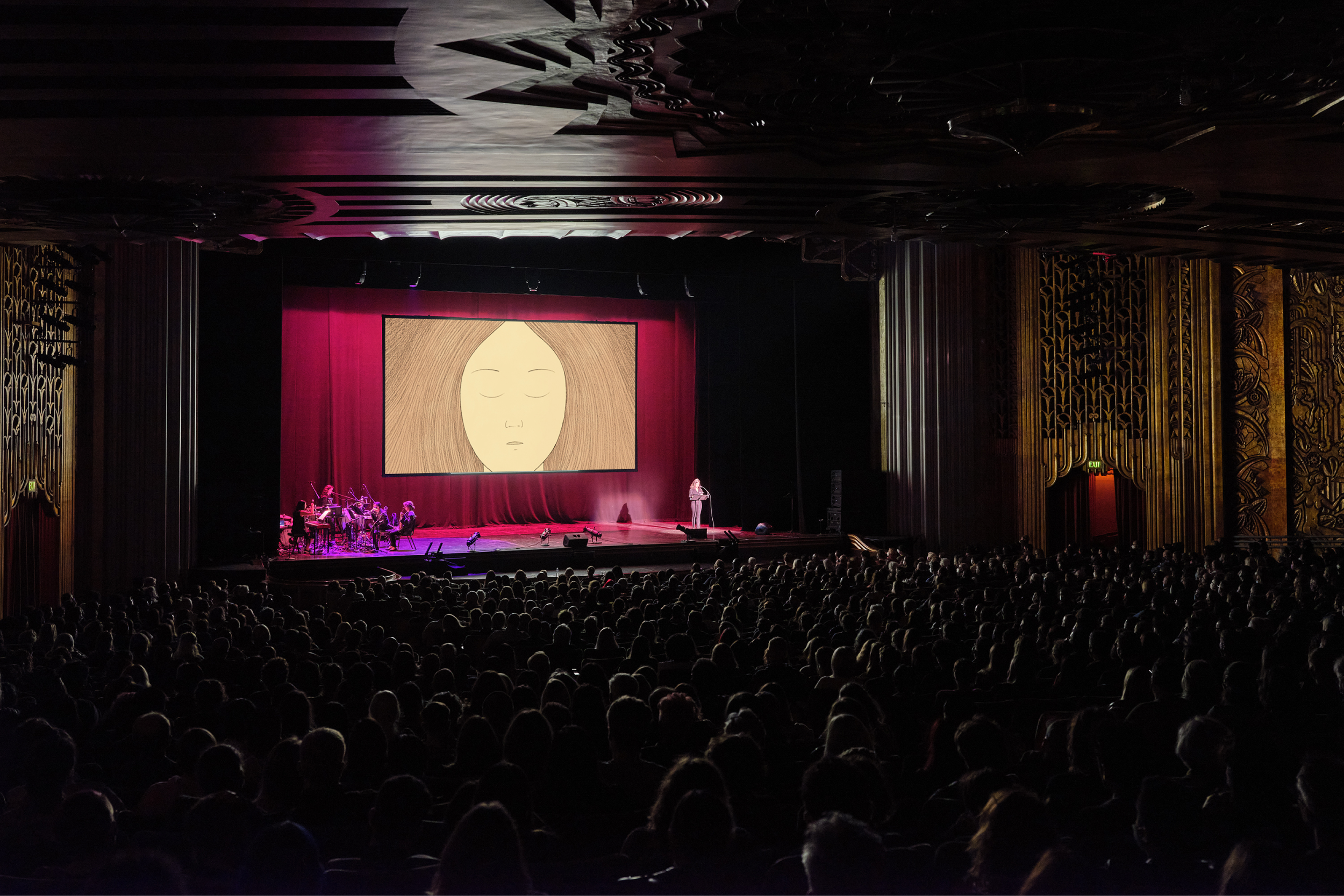 Ingrid Rojas Contreras performs on stage with Magik*Magik Orchestra. Behind her is a screen with artwork of a woman with her eyes closed.