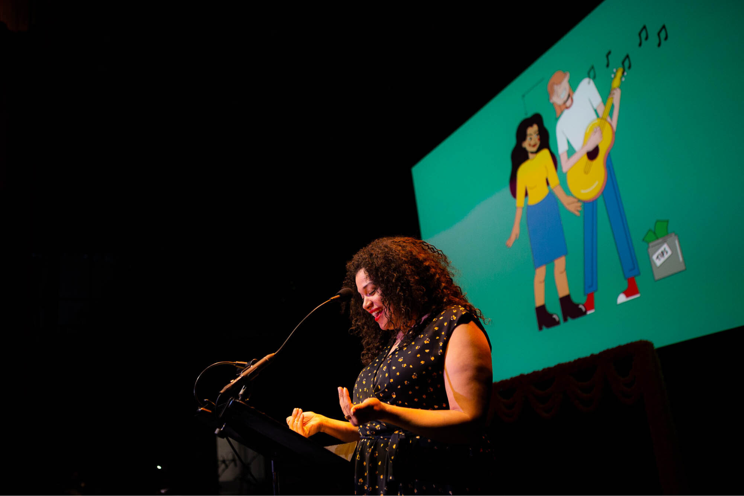 Michelle Buteau performs on stage in front of a screen with a drawing of a couple.