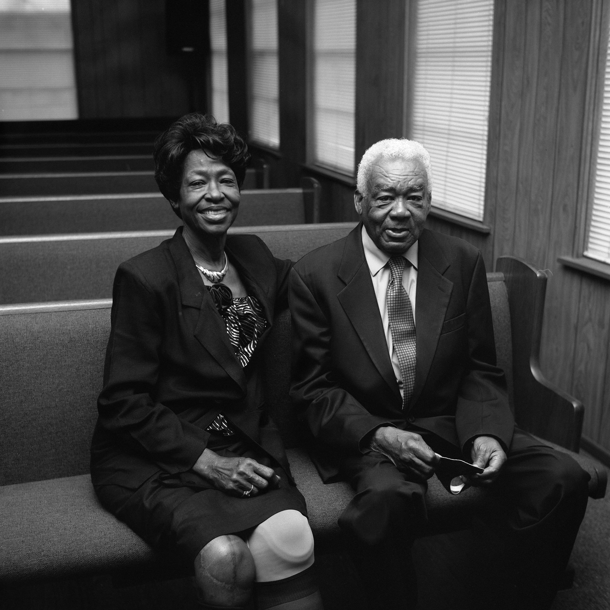 Image description: a black-and-white photograph of an elderly African American man (wearing a suit and tie) and woman (wearing a blazer, blouse, and skirt) seated in a church pew, smiling at the camera. Art by Rahim Fortune.