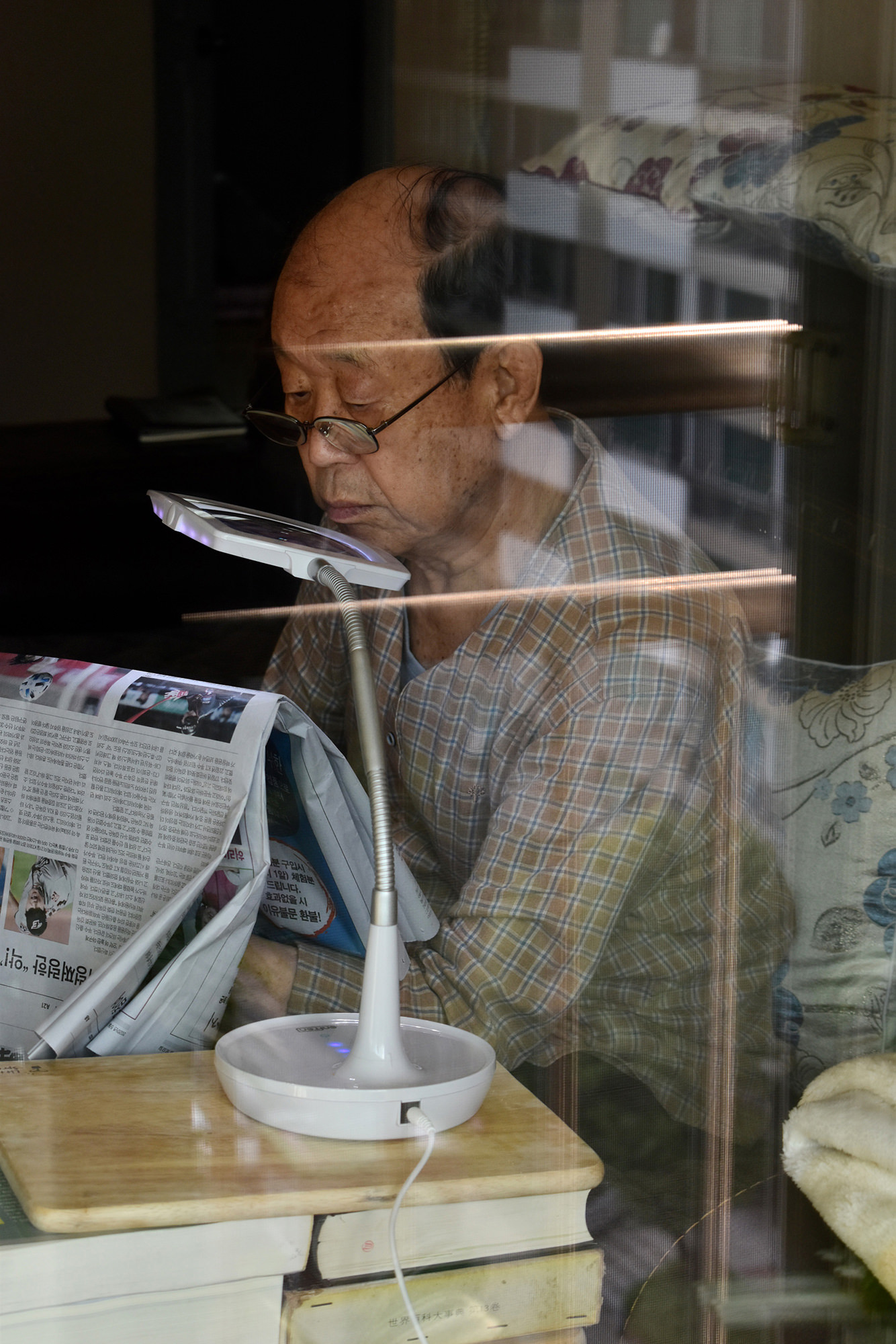 Image description: the artist’s Asian grandfather reading a newspaper, unaware of the camera and photographed through glass, which shows reflections. Photo by Annie Son, 18.