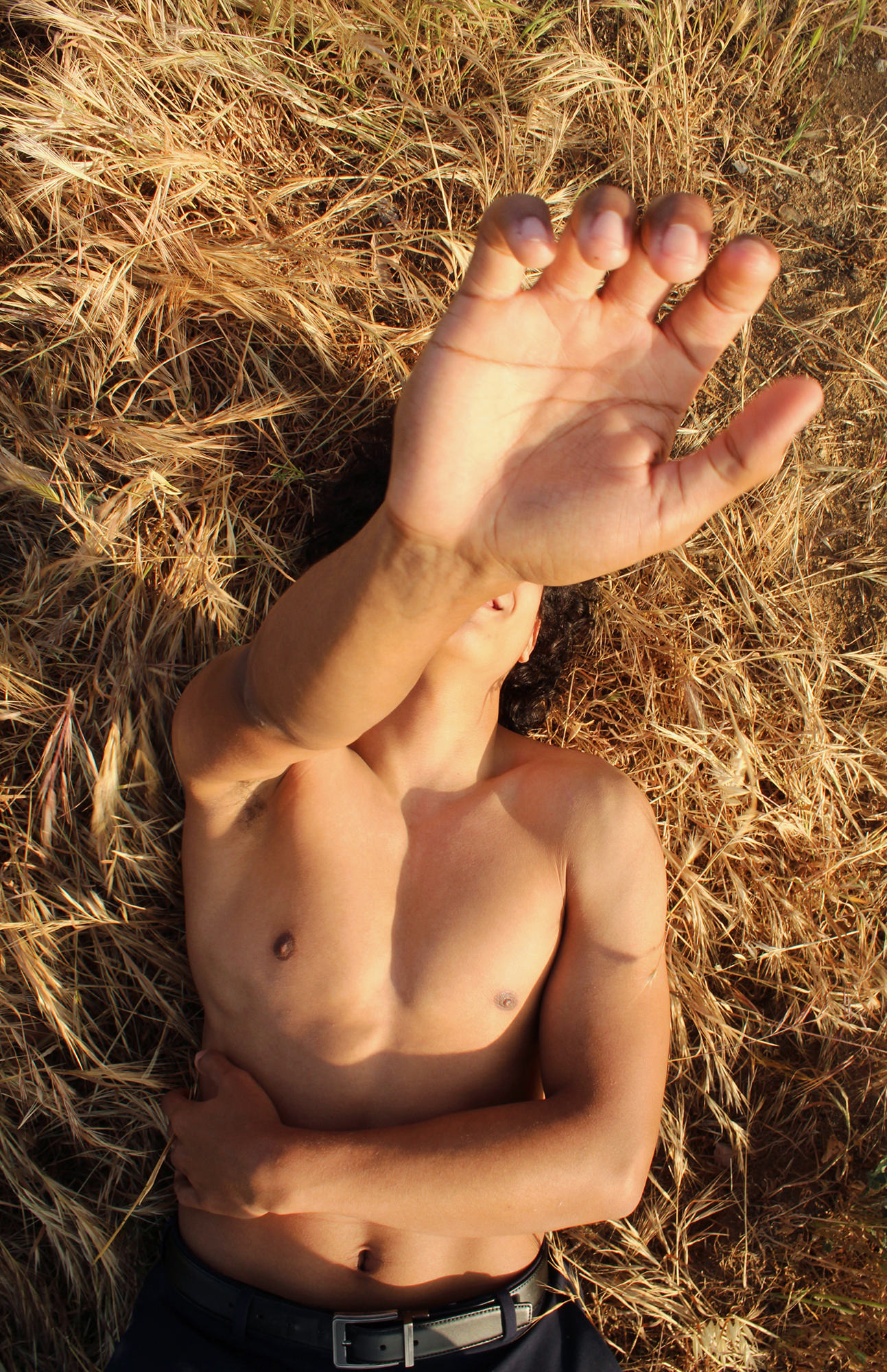 Image description: a portrait of the artist’s partner, a young man lying in straw grass without his shirt, hand up in the air obstructing his face to the camera. Photo by Valeria Hernandez, 17.