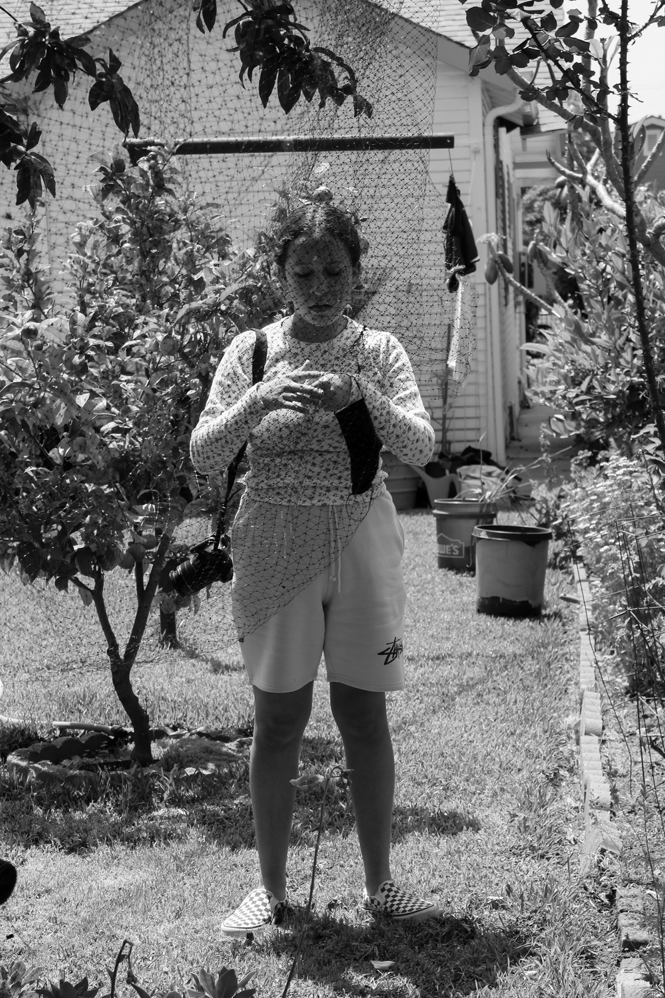 Image description: a black-and-white photograph of the artist’s older sister looking down at her hands in a backyard behind a net. Photo by Ixchel Cruz, 15.