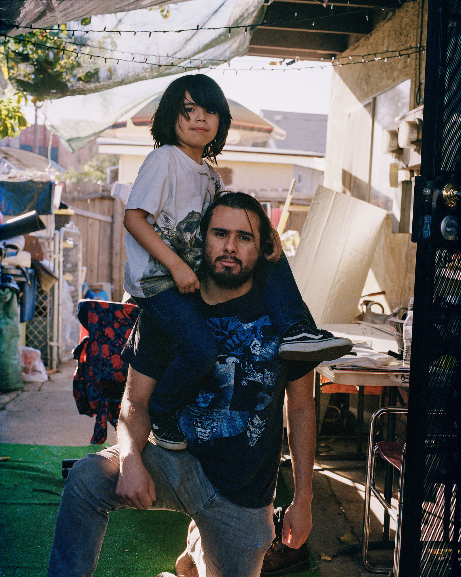 Image description: a photograph of a young boy resting on his father’s shoulders, who is kneeling, outside of their front door. Photo by Star Montana.