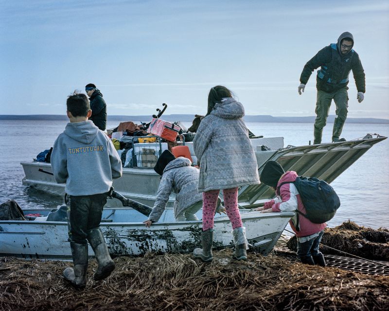 Children climb onto a small boat on a river, while a second boat, loaded with belongings, is behind it.