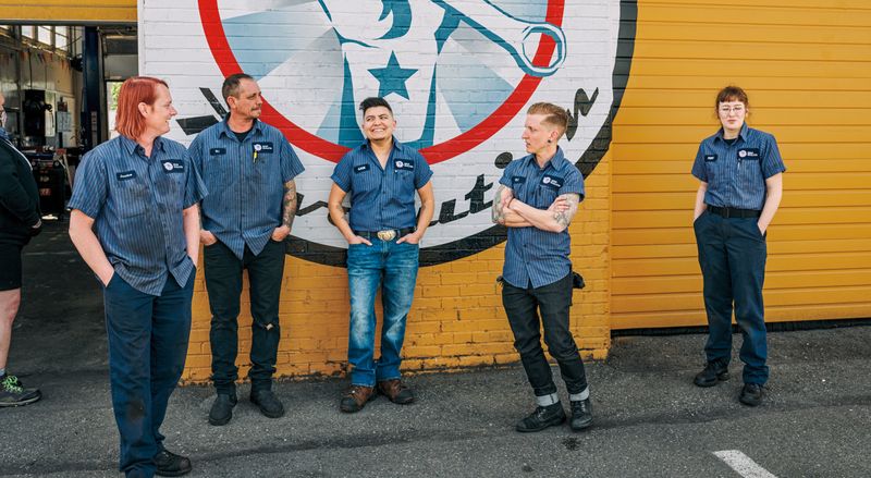 Five mechanics in matching uniforms stand in front of a wall with a mural of a hand holding a wrench.