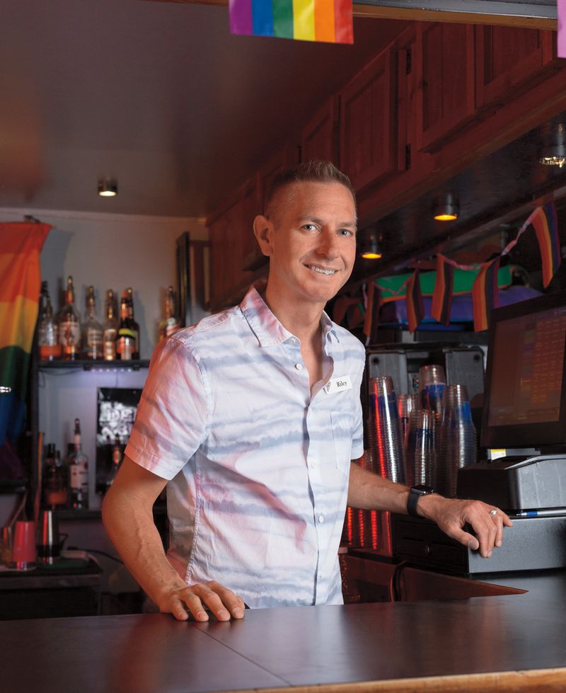 A smiling man is standing behind a bar with rainbow flags in the background.