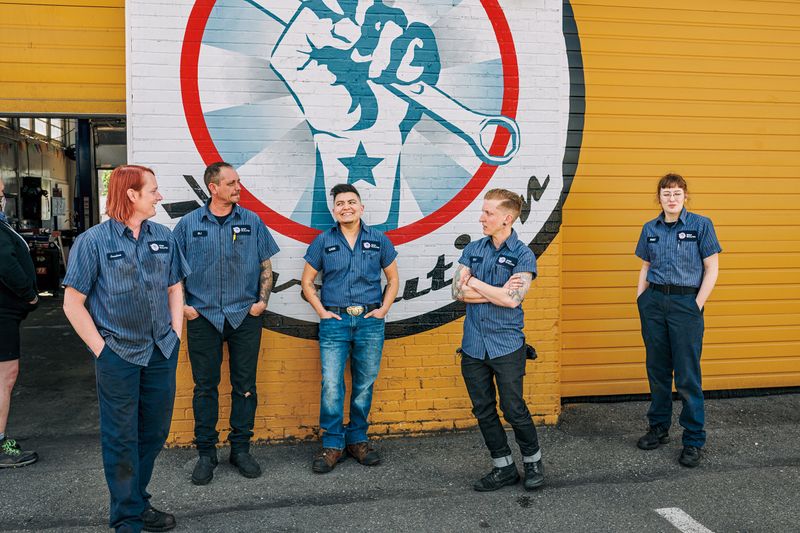 Five mechanics in matching uniforms stand in front of a wall with a mural of a hand holding a wrench.