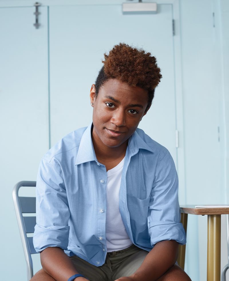 A person in a blue shirt and shorts is sitting on a chair, looking at the viewer.