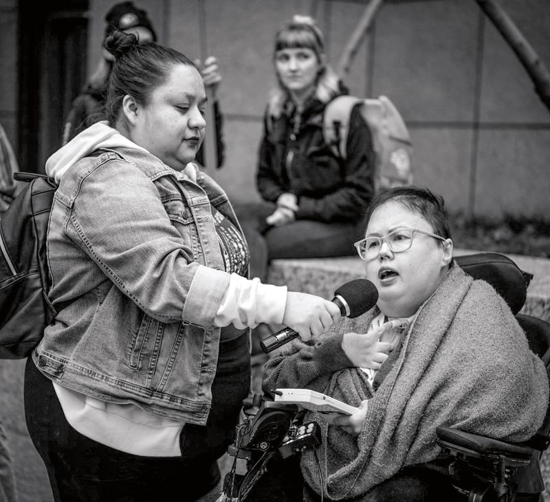 A woman in a wheelchair speaks into a microphone held by a woman standing by her side.