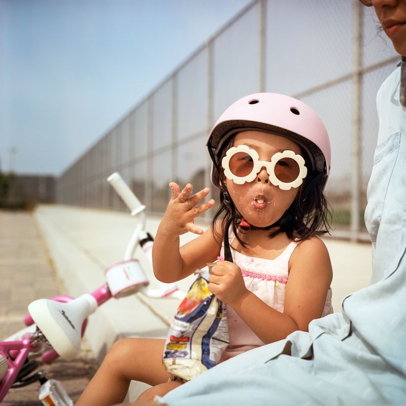 Una niña con un casco rosado y gafas de sol blancas come un refrigerio.