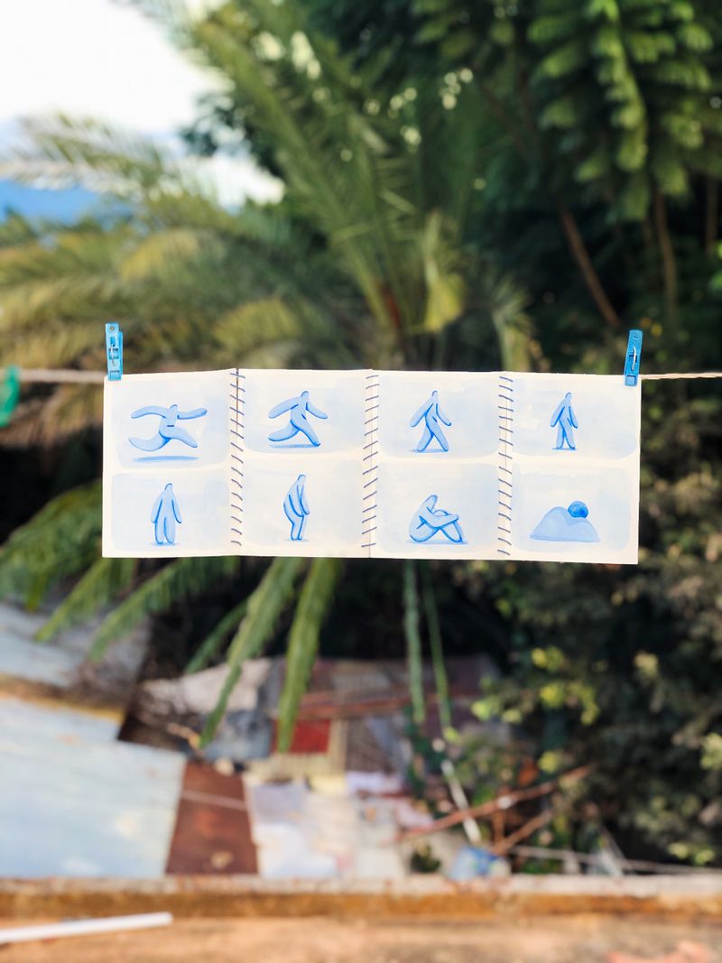 A photo of trees in the background and a clothesline holding illustrations in the foreground. There are eight illustrations sewed together of a blue cartoon-like figure in various positions.