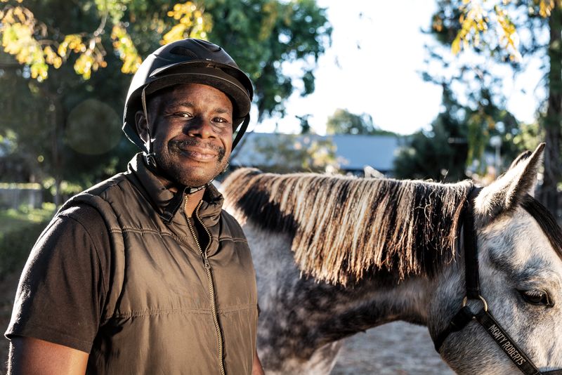 A man in a riding helmet stands smiling beside a horse.