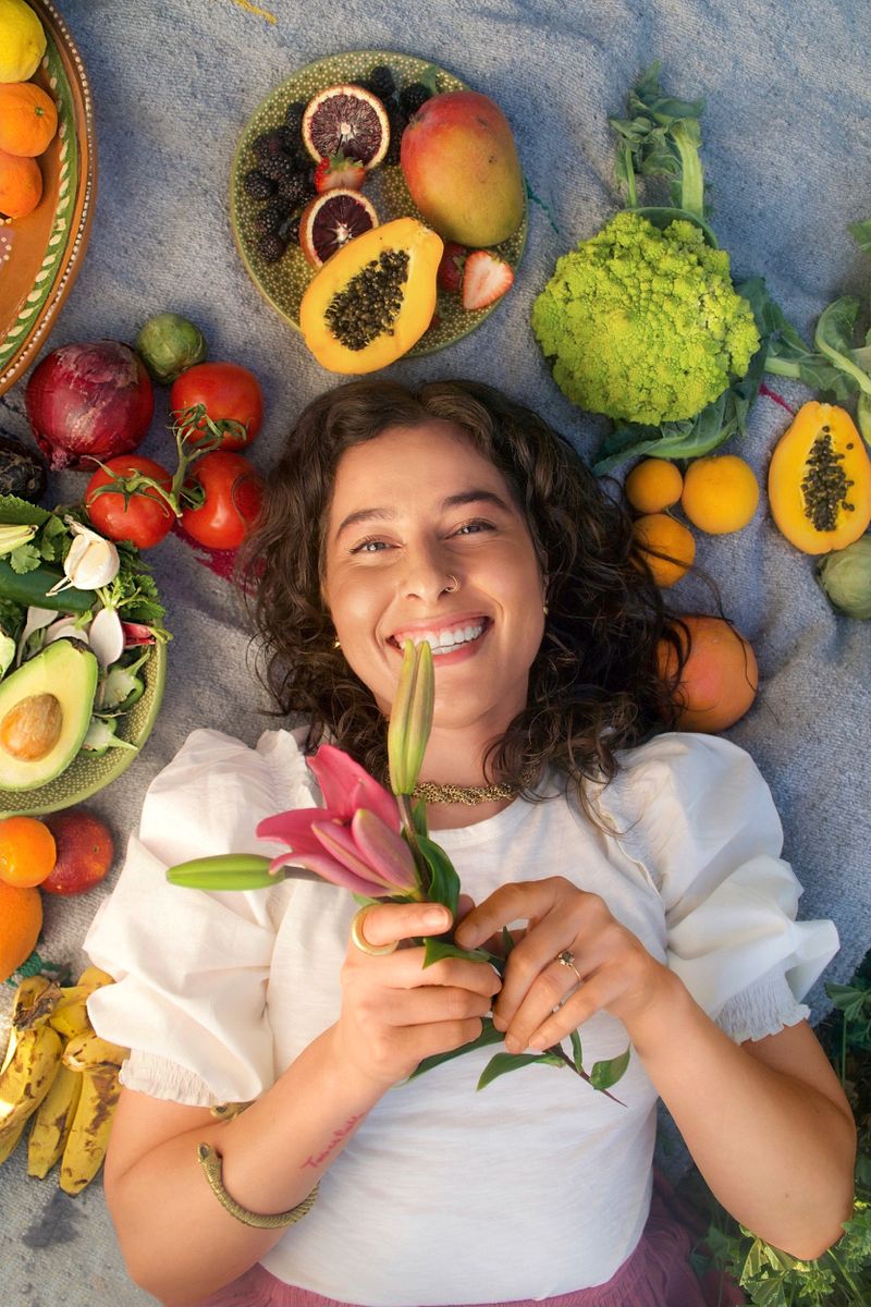 Una mujer con una blusa blanca acostada de espaldas, rodeada de frutas y verduras de colores brillantes, sostiene una flor rosada.