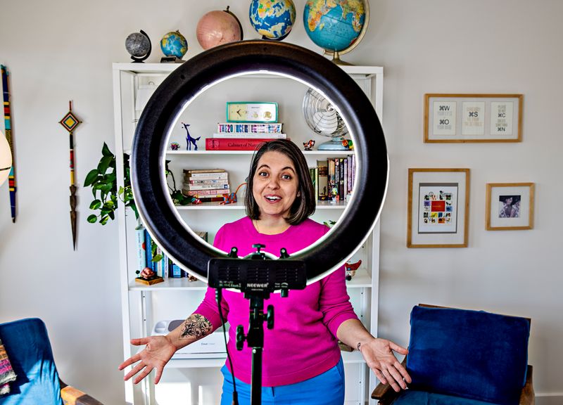 A woman stands, smiling through a ring light.