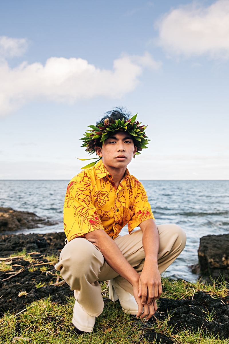 Boy sitting on his haunches in front of the ocean, wearing a crown of leaves.