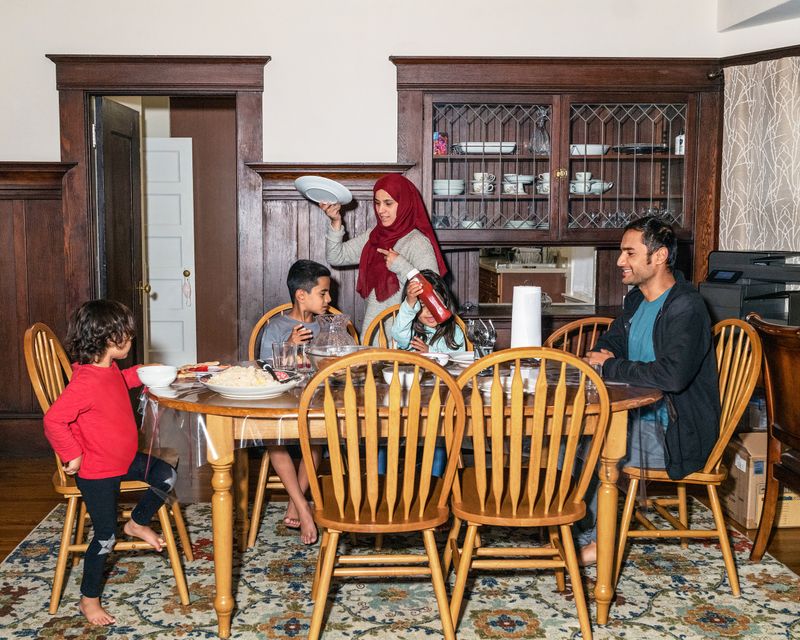 A man and three children sit at a dinner table while a woman walks behind them carrying a plate.