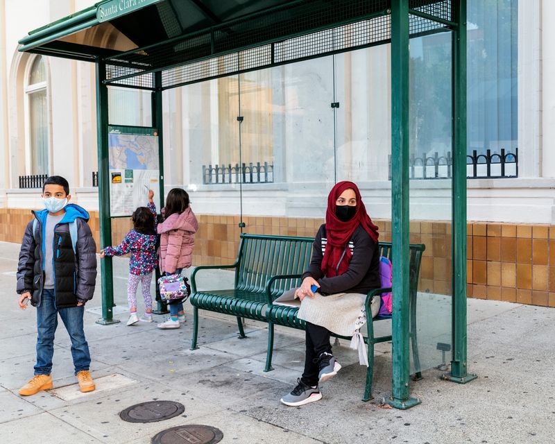 A woman sits on a bench at a bus stop with her three children, who are standing.