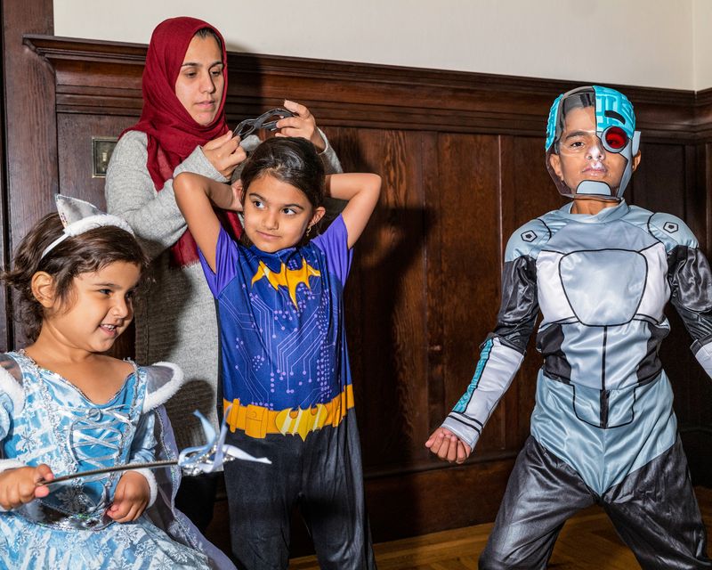 Three children stand, dressed up for Halloween, while a woman helps the one put on a mask.