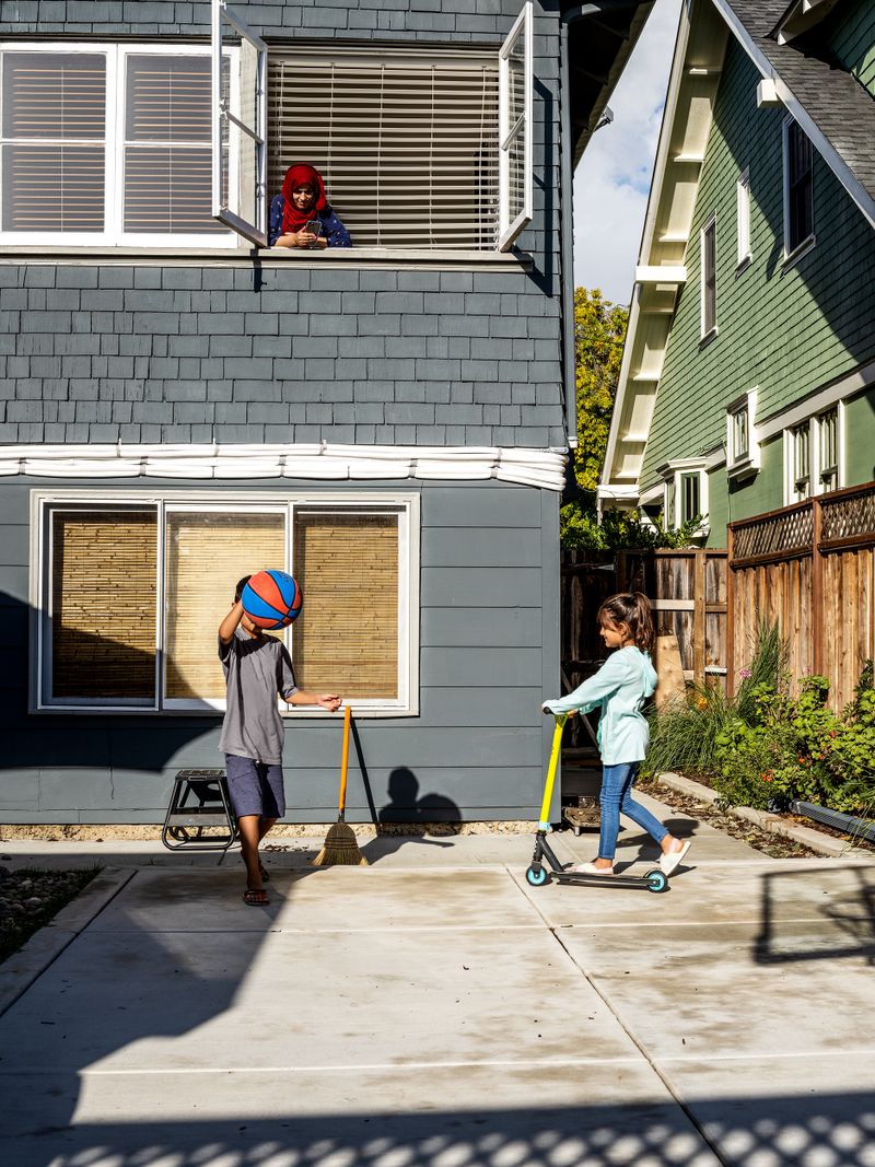 A woman leans out a second-floor window to watch her two children playing below, one with a basketball and one on a scooter.